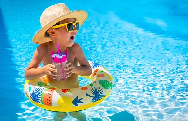 A boy in the pool wearing a hat, goggles, float, holding a drink