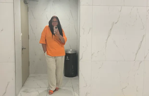 Woman in orange blouse and beige pants in front of a bathroom mirror