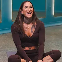 A woman wearing brown workout attire, she is sitting and smiling, she has long dark hair
