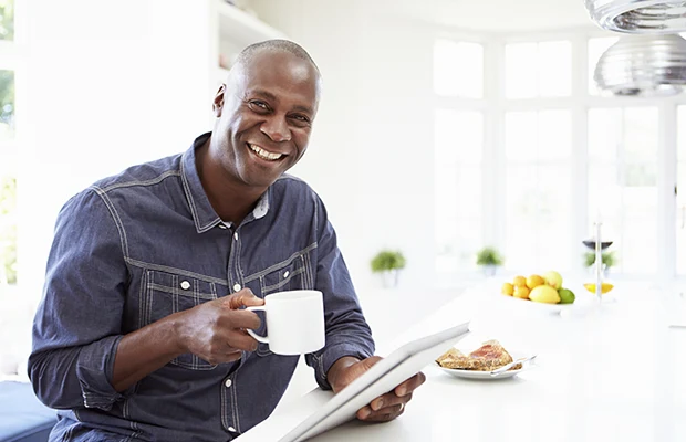 A middle aged man holding a cup, sitting in a kitchen, and smiling at the camera. There's a plate of bread next to him.