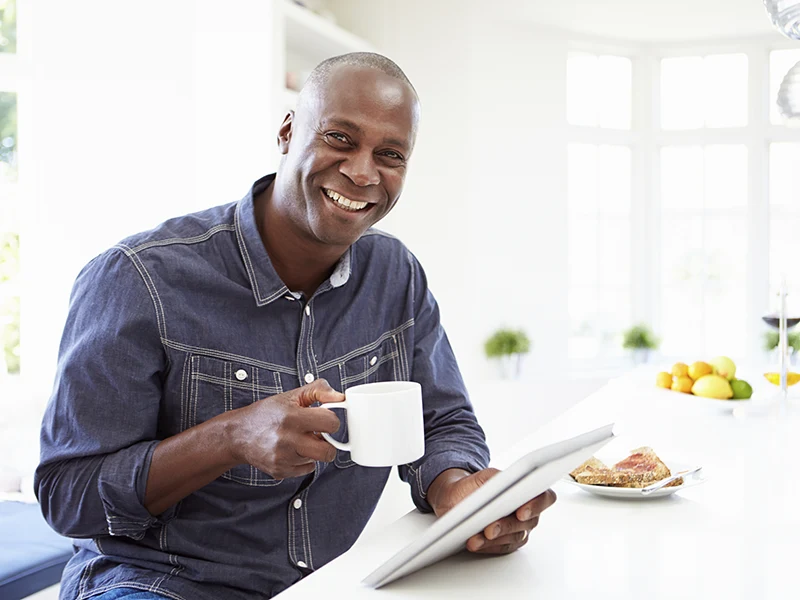 A middle aged man holding a cup, sitting in a kitchen, and smiling at the camera. There's a plate of bread next to him.