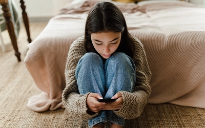 girl sitting on the floor of her room in front of the bed, while using her cell phone