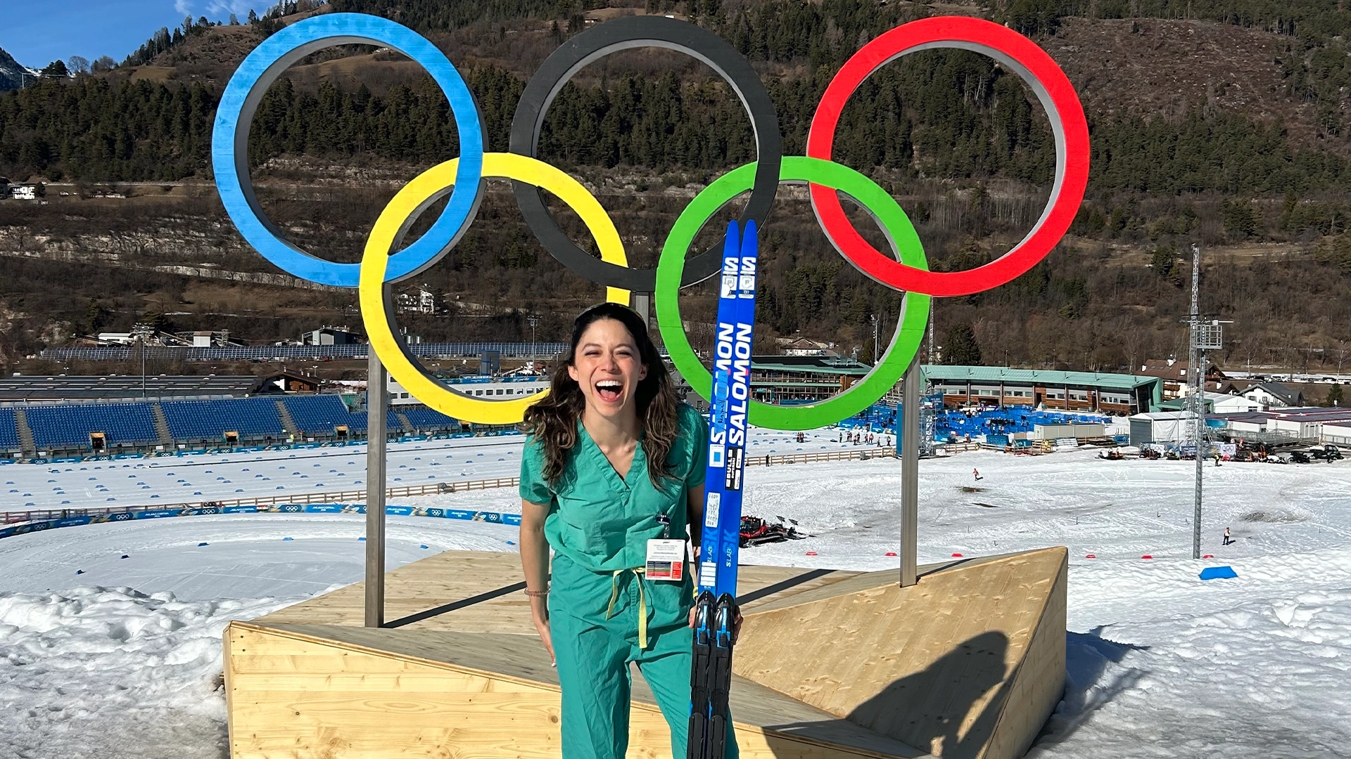 Woman with skis next to a mountain and the Olympic Games logo.