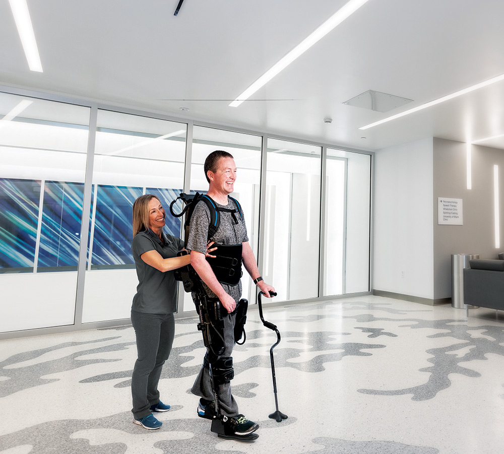 A male wearing an Exoskeleton device and a cane to help him walk, a health care provider stands behind him helping him walk