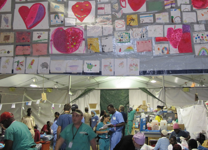 Physicians in a field hospital providing care to numerous people, you see hand drawings on the wall