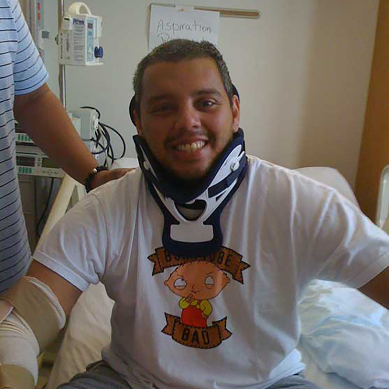 Carlos Castillo sitting up on a hospital brace, he is smiling and wearing a neck brace