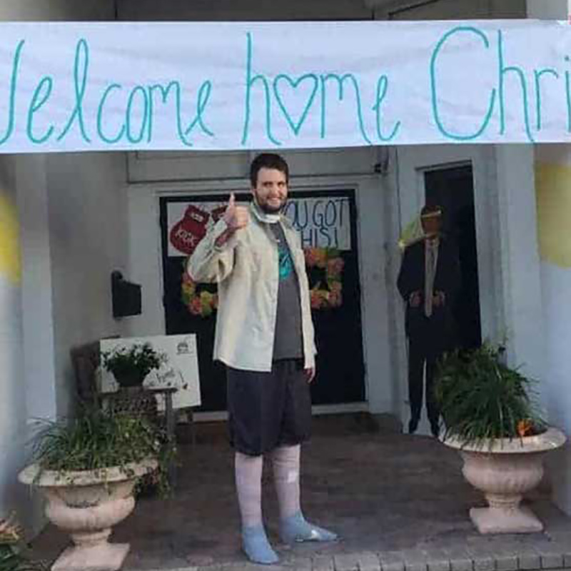 Chris Rotella standing in front of a doorway of a home with his thumb up, there's a sign above him that says welcome home Chris