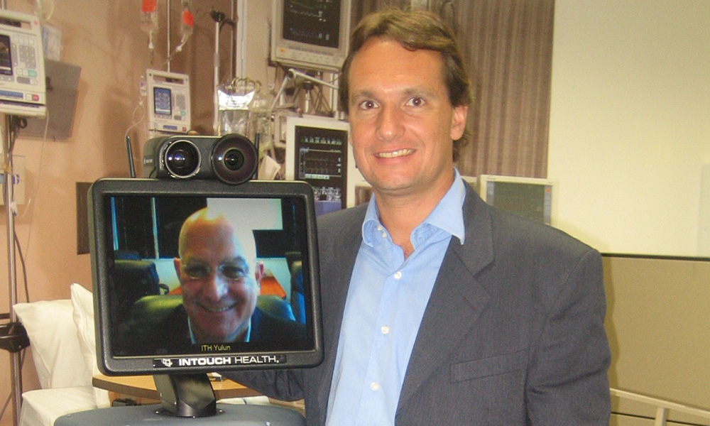 A male wearing a gray suit jacket and blue shirt standing next to a robot that has a male within the screen