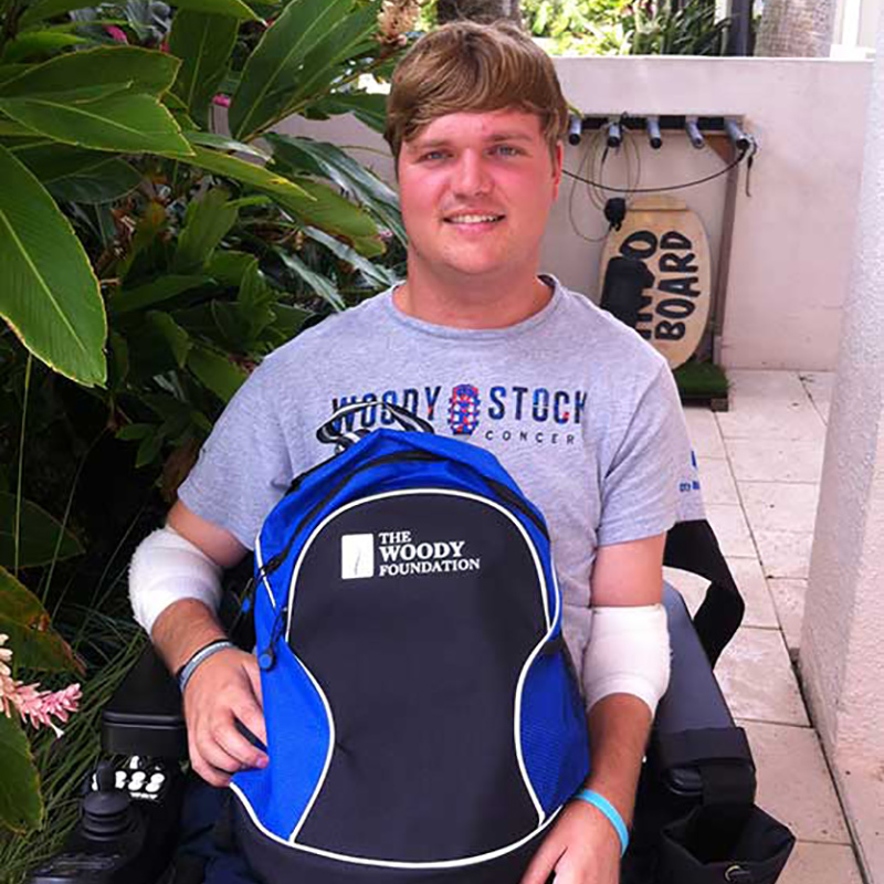 Woody sitting in his wheelchair, he's holding a backpack that says The Woody Foundation