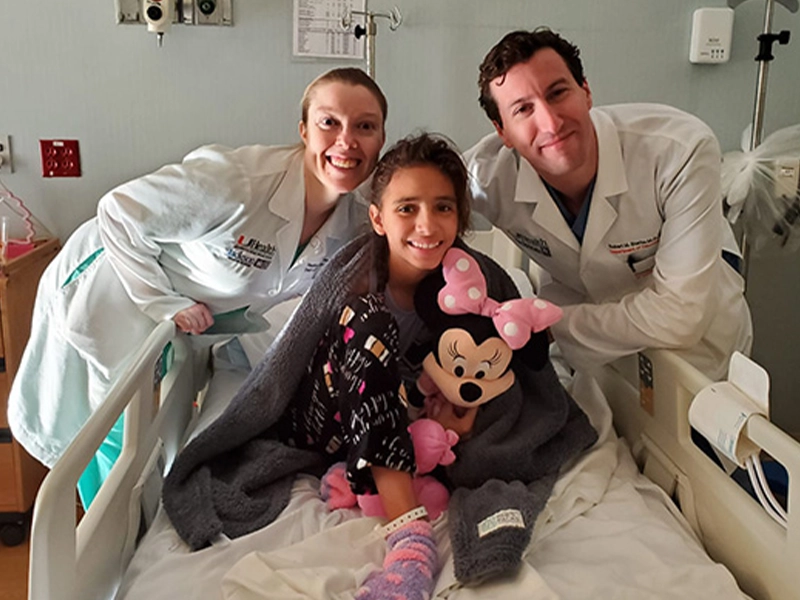 Sasha Gadea sitting up on a hospital bed with her two doctors behind her smiling for the camera