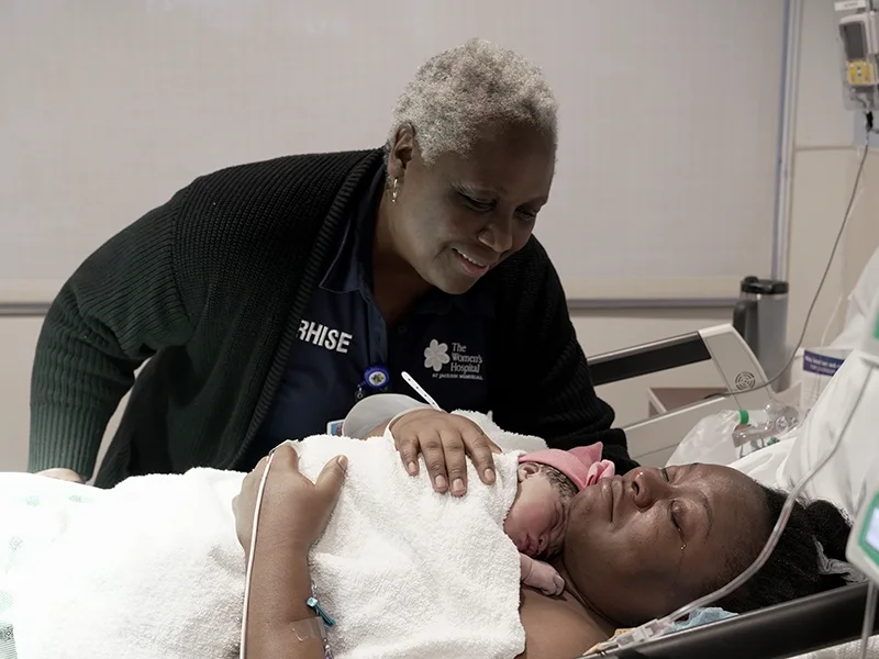 A medical professional smiling as she looks over a patient who just gave birth, the patient is holding her baby on her chest