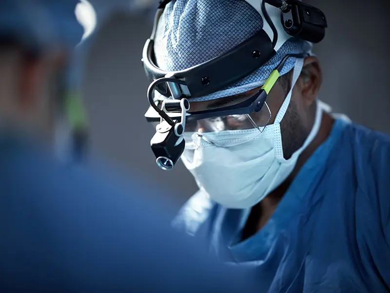 A closeup of a surgeon, they have on a light around their forehead, glasses, and are wearing a face mask