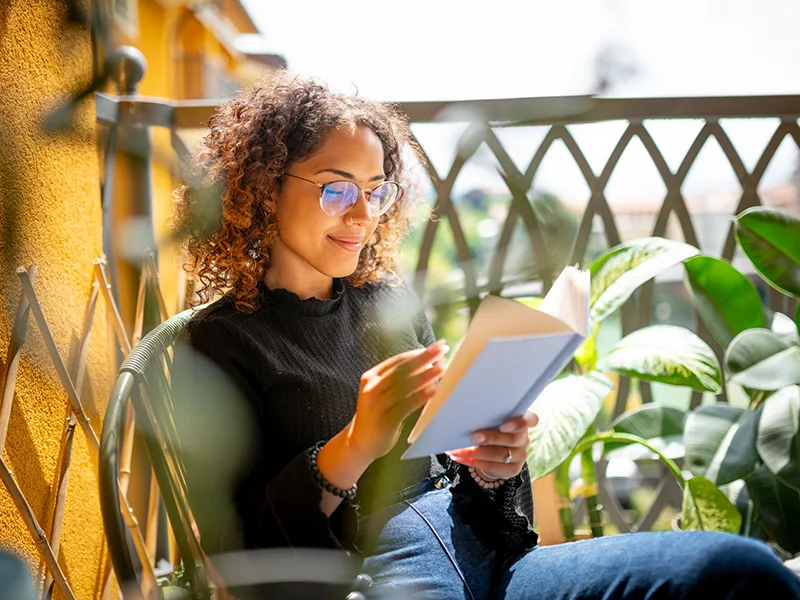 A woman sitting outdoors reading a book, she has short curly hair and is wearing glasses