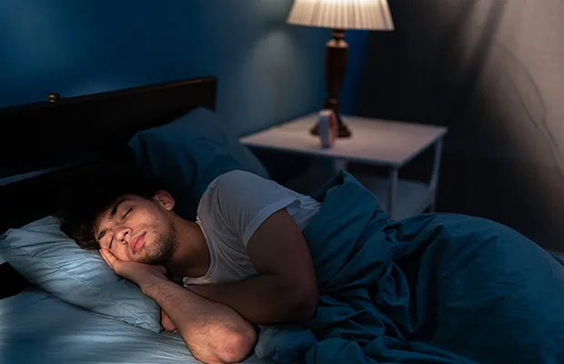 A man sleeping in his bed, behind him there's a lamp turned on