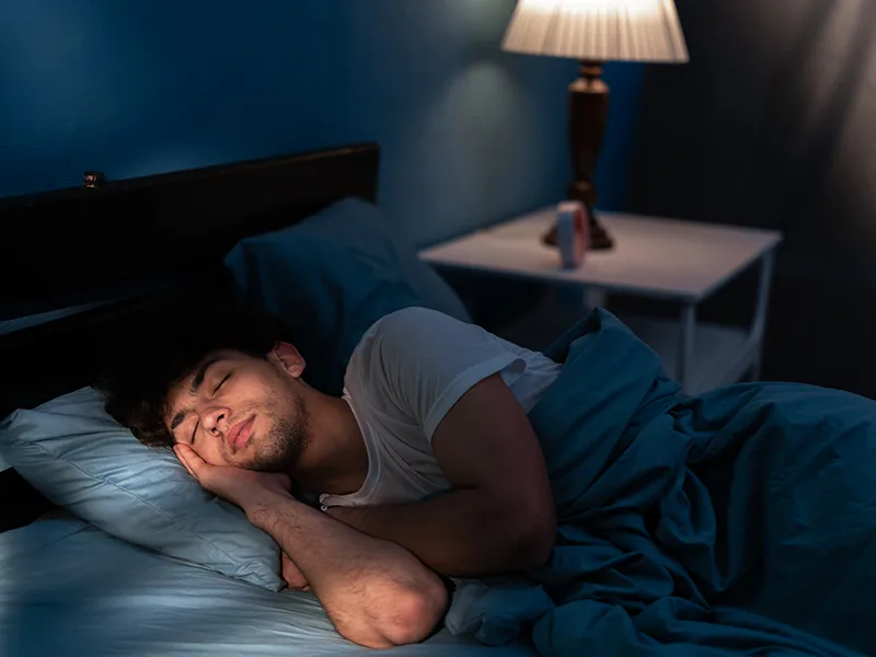 A man sleeping in his bed, behind him there's a lamp turned on