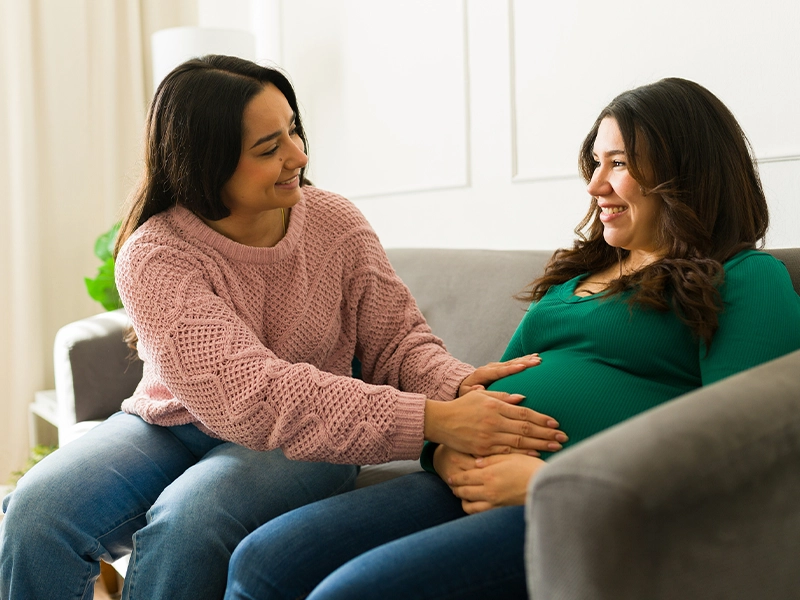 A woman touching a pregnant woman's vulture while sitting on a sofa and smiling