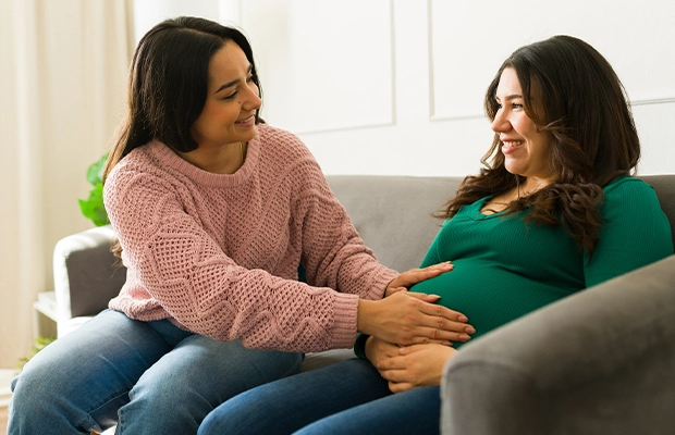 A woman touching a pregnant woman's vulture while sitting on a sofa and smiling