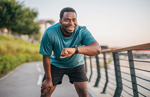 A man outdoors, he is leaning forward and checking his watch, it looks like he is on a run