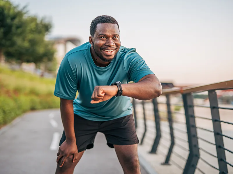 A man outdoors, he is leaning forward and checking his watch, it looks like he is on a run