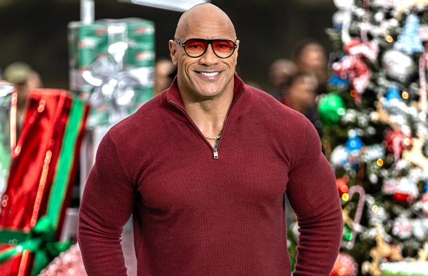 A photograph of The Rock taken during an event; he is wearing a deep red sweater and glasses of the same color.