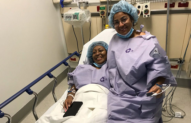 Two women wearing hospital gowns, they are seated on a medical bed and smiling at the camera