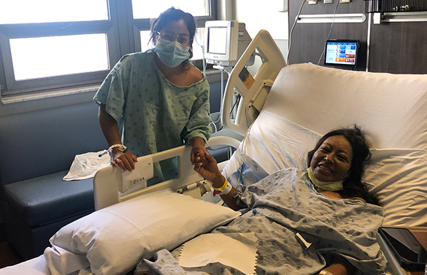 Two women in a hospital room, both wear gowns, one is standing holding her friends hand while the other is laying on a medical bed
