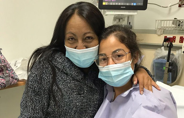 To woman in a hospital room, both are wearing masks and looking at the camera, one wears a hospital gown and another a gray coat