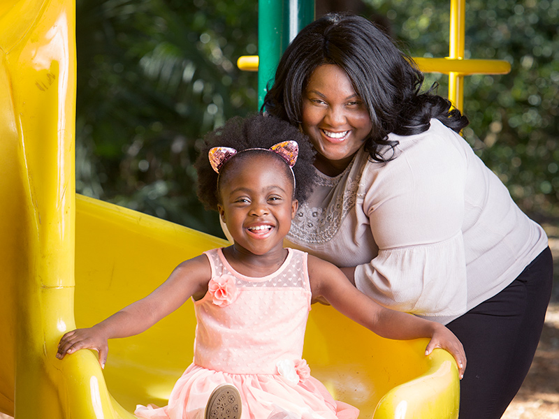 Tanisha at the bottom of a slide with her mom standing behind her, they are both smiling at the camera