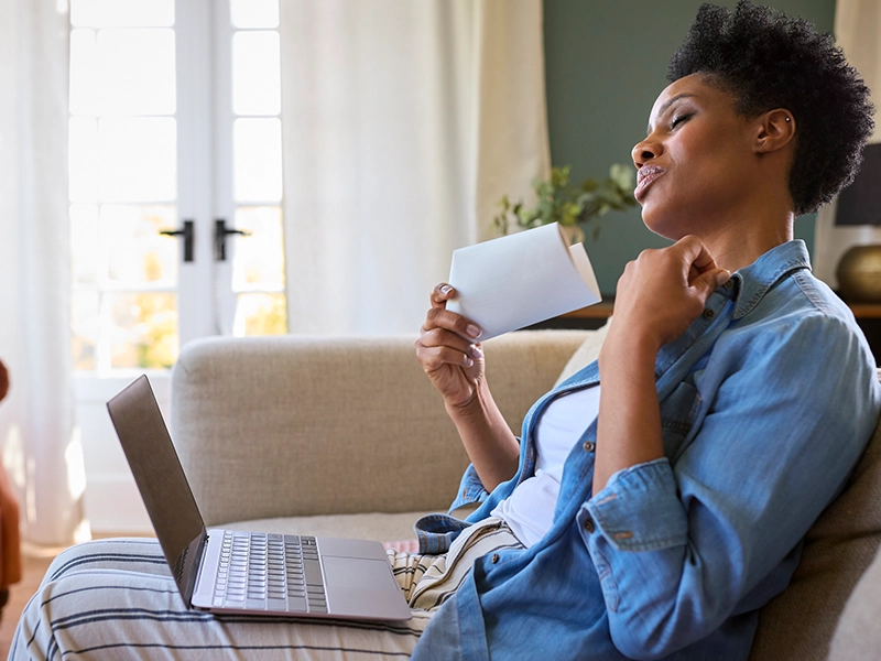 woman sitting on the couch with a computer on her legs fanning herself with a piece of paper