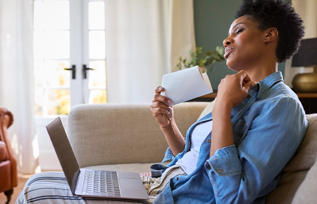 woman sitting on the couch with a computer on her legs fanning herself with a piece of paper