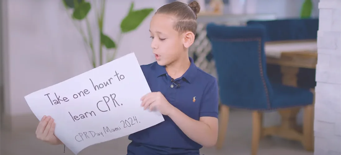 A young boy holding a sign that reads Take on hour to learn CPR. CPRDayMiami2024