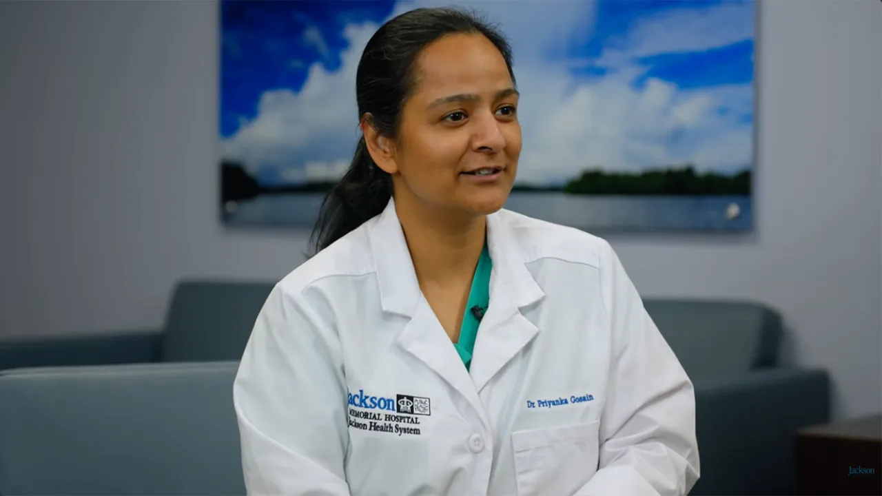 A medical professional looking to her left, she has on a white coat and is sitting on a couch