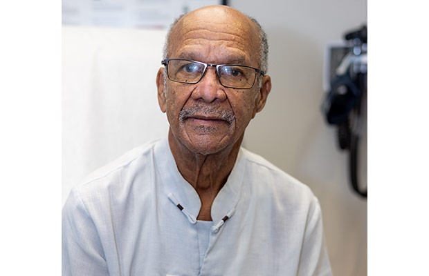 A closeup of an older man looking at the camera, he is sitting inside a medical patient room