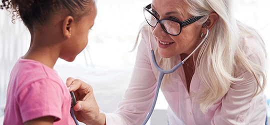 Female pediatrician with white hair and glasses using a stethoscope to check a young girl's heartbeat.