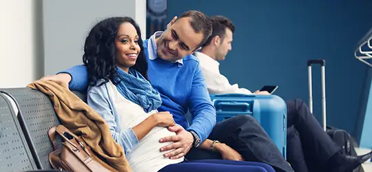 Pregnant woman smiling, her partner touches her belly, they smile as they sit at an airport lounge