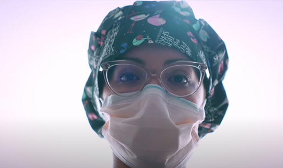 A closeup of a medical professional, she has glasses on, a face mask, and a nursing cap