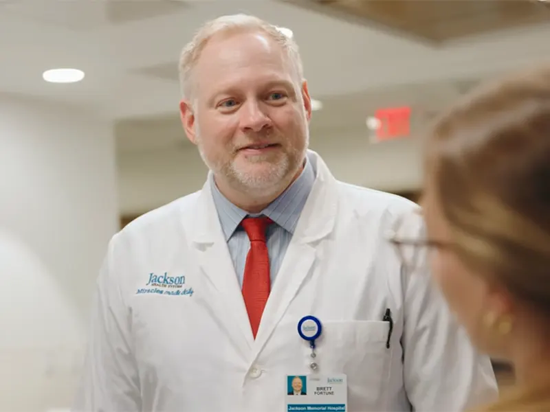 A medical professional in a medical hallway speaking to a colleague, he has on a medical coat, blue shirt, and red tie