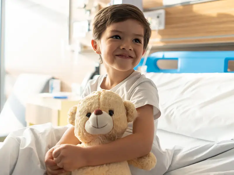 A young boy on a hospital bed smiling, he holds a teddy bear