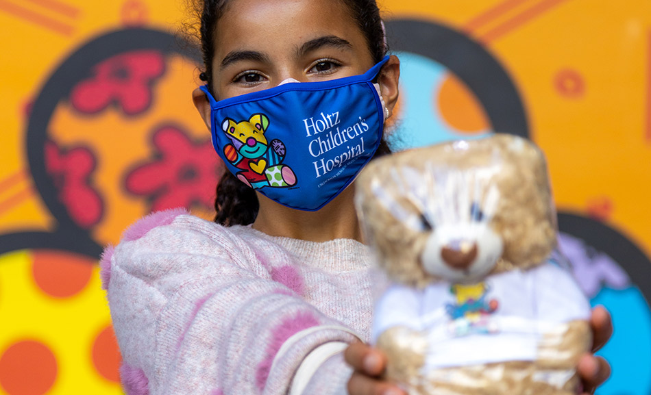 Young girl holding a teddy bear, she wears a face mask that reads Holtz Children's Hospital