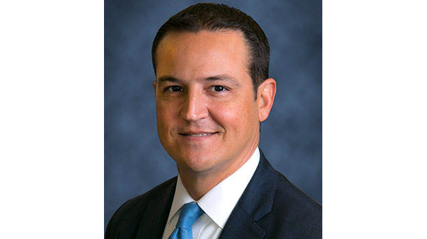 A headshot of a man, he has short black hair and is wearing a navy suit and tie