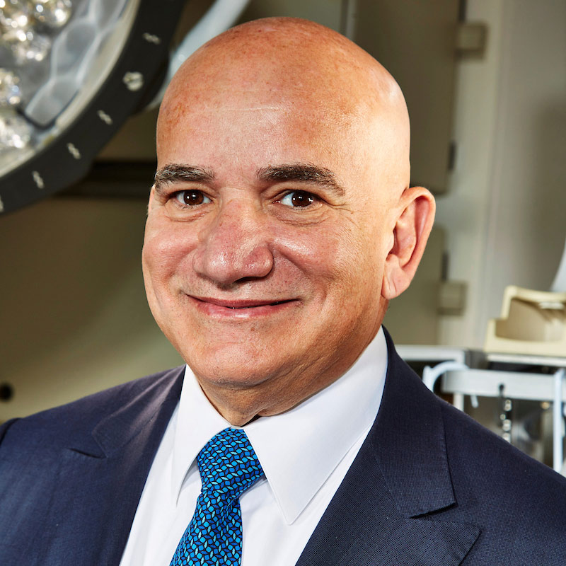 A closeup of a man who smiles at the camera, he is wearing a navy blue suit jacket, a white collar shirt and a blue tie