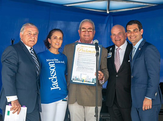 A group of four man and one woman smiling at the camera, they all stand next to each other, the man in the center of the photo holds a proclamation sign