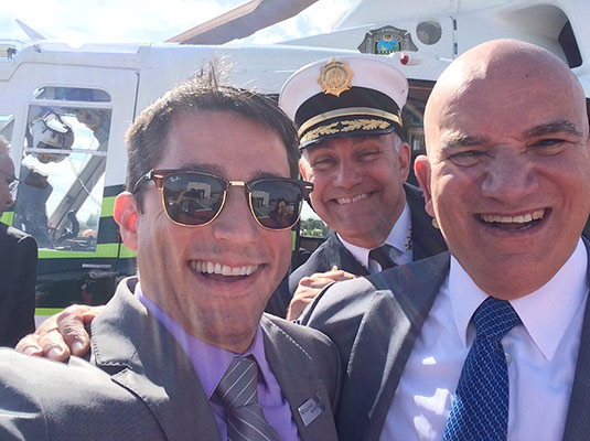 Three men smiling, they are outdoors and standing in front of a helicopter