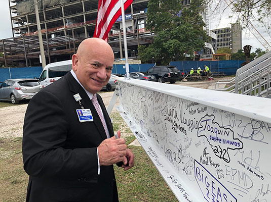 A man in a suit and tie outdoors smiling at the camera, he stands in front of a white wall full of signatures