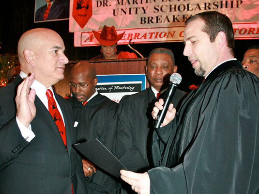 A man in a suit and tie with his right hand up, another man speaks into a microphone, there are people standing behind them