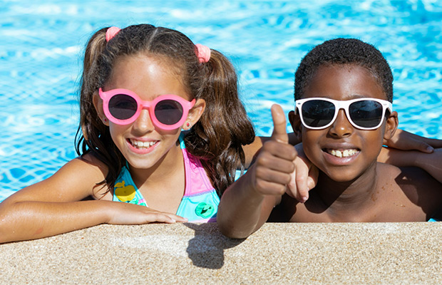 Two young children smiling at the camera, they are in the pool, both are wearing sunglasses and one child is giving a thumbs up
