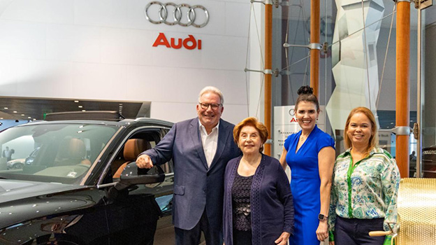 Four adults, one man and three woman, standing next to a black Audi vehicle within a car dealership, they are all smiling at the camera