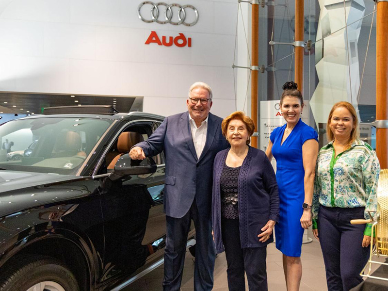 Four adults, one man and three woman, standing next to a black Audi vehicle within a car dealership, they are all smiling at the camera