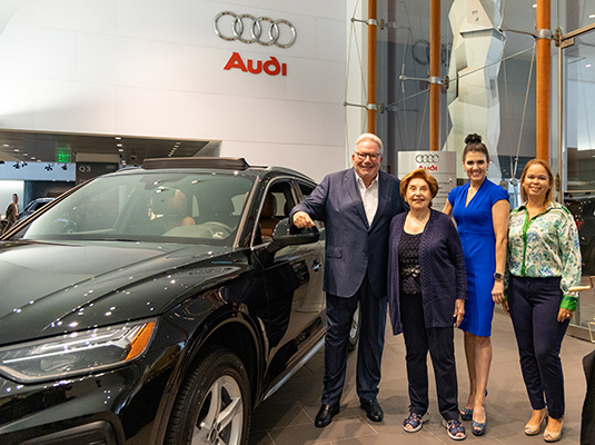 Four adults, one man and three woman, standing next to a black Audi vehicle within a car dealership, they are all smiling at the camera