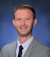 Headshot of Gradon, he is wearing a gray suit jacket, a dark tie, and a light violet collar shirt